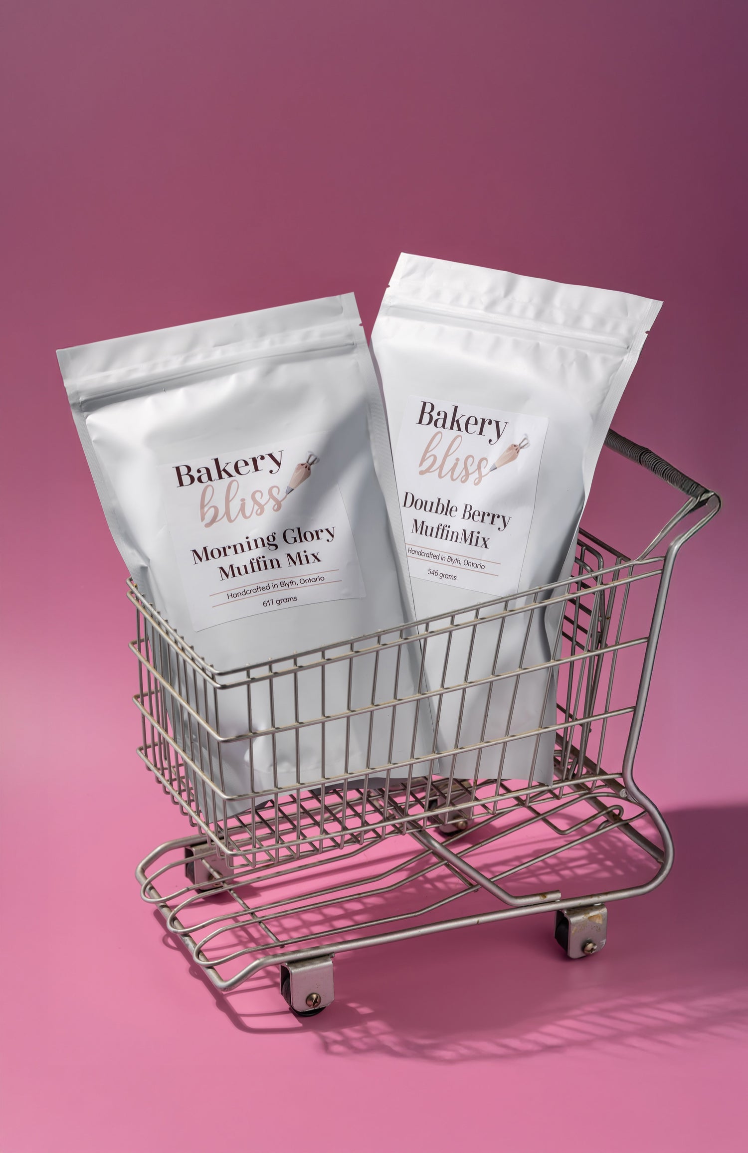 A miniature shopping carts containing 2 Bakery Bliss Muffin Mixes on a pink background