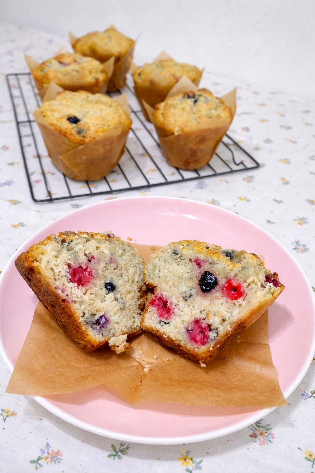 Sliced berry muffin on a pink plate with whole muffins on a cooling rack in the background.
