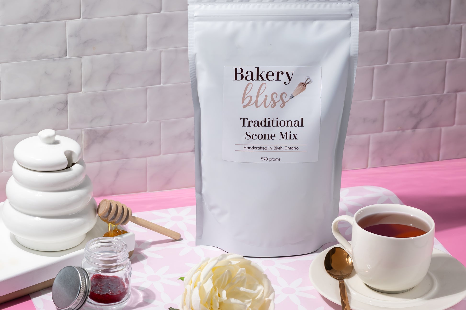 Bakery bliss Traditional Scone Mix bag with a cup of tea, honey jar, and scone on a pink and white checkered tablecloth.