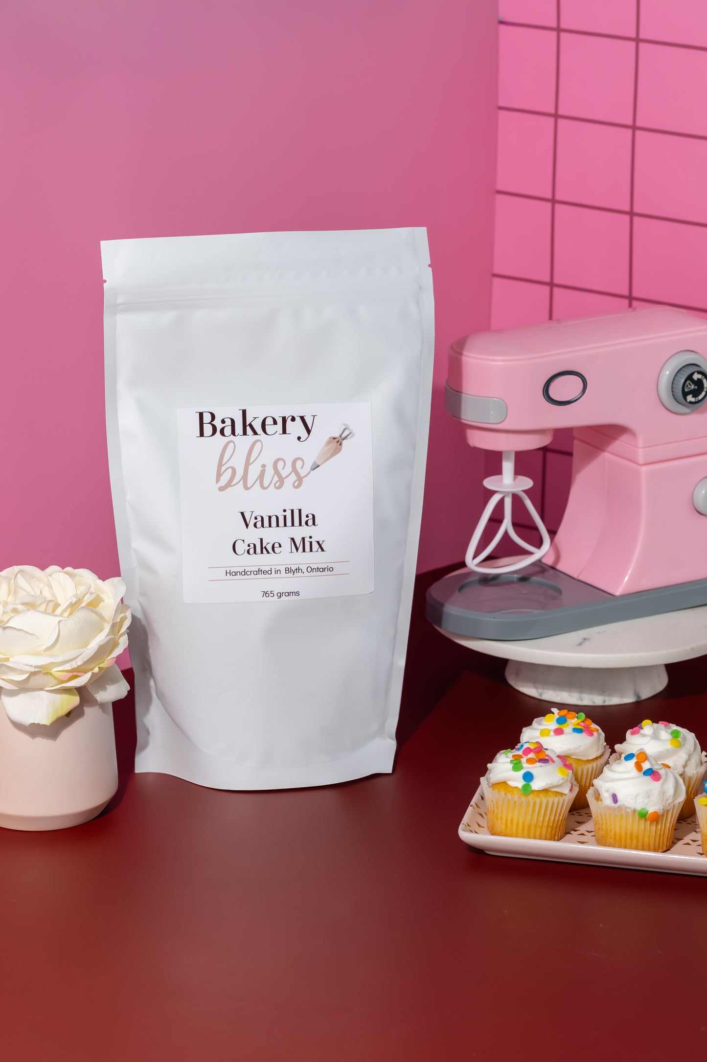 Bakery Bliss vanilla cake mix package with a pink mixer and cupcakes on a red surface.