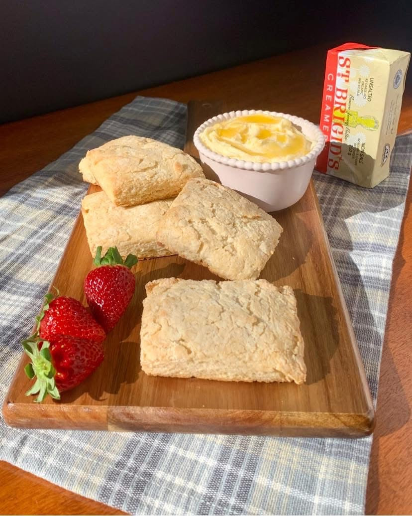 Wooden tray with scones, honey butter, and strawberries on a checkered tablecloth.