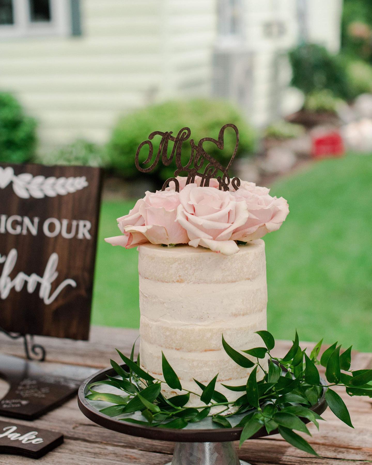 Small wedding cake with pink flowers and a 'Mrs & Mr” topper on a wooden table outdoors.